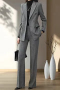 A woman models a gray double-breasted blazer and matching wide-leg trousers set, styled with a black turtleneck, black shoes, and a black handbag in a minimalist room with warm lighting.