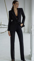 A woman modeling a black tailored pantsuit with a structured blazer and straight-leg trousers in a bright white interior setting.
