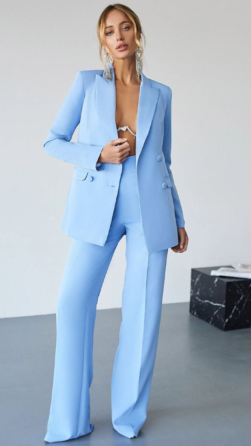 A woman models a light blue tailored pantsuit with a single-breasted blazer and wide-leg trousers in a minimalist studio setting.