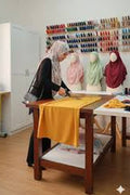 Person working at a table with a yellow cloth in a room with a display of headscarves on the wall.