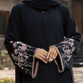 A person wearing a black abaya with long sleeves featuring pink and white floral embroidery and fringe trim, paired with a matching black headscarf. The background is a softly blurred outdoor setting.