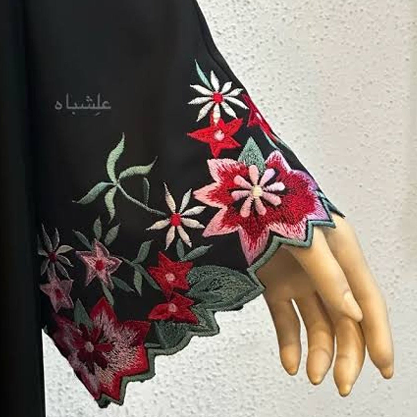 A close-up of a black abaya or long dress sleeve with scalloped edges and colorful floral embroidery in red, pink, white, and green, displayed on a mannequin hand against a light textured wall.