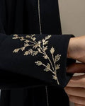 Close-up of a black garment sleeve with delicate gold floral embroidery, held by a hand against a neutral background.