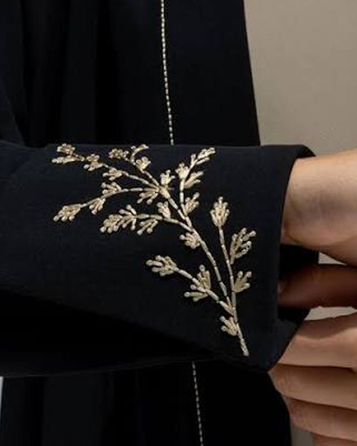 Close-up of a black garment sleeve with delicate gold floral embroidery, held by a hand against a neutral background.