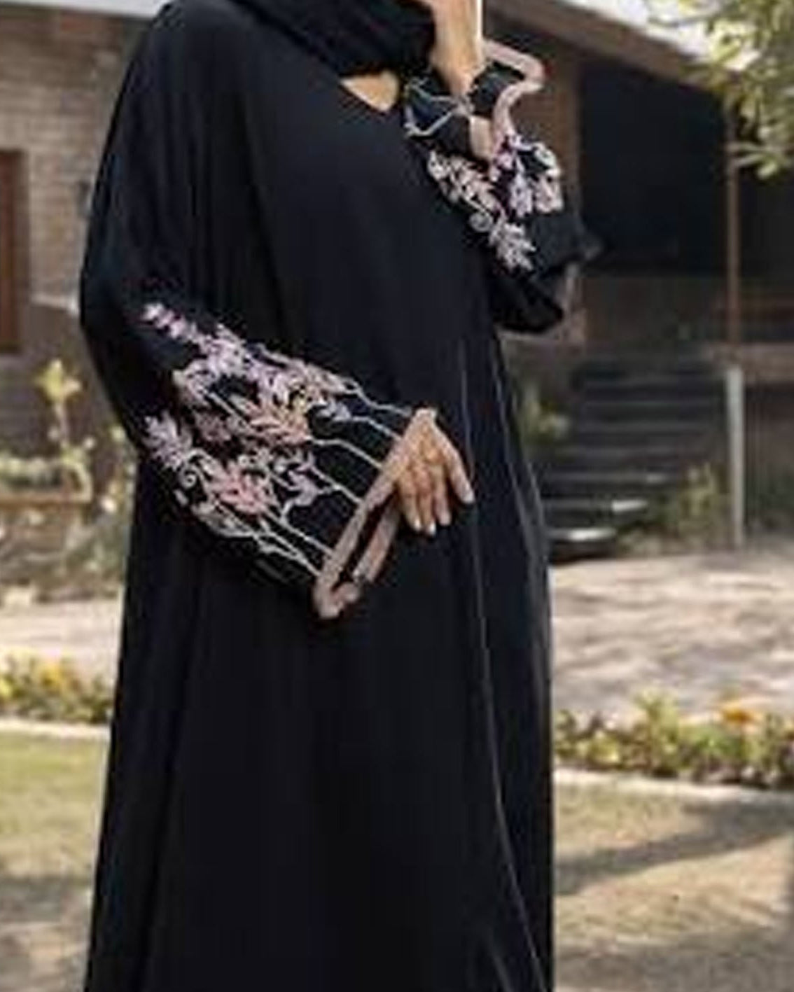 A woman wearing a black abaya with pink floral embroidery on the sleeves, paired with a matching black hijab, standing outdoors in front of a house.