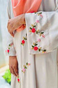 A close-up of a person wearing a light beige or cream-colored long-sleeved garment with colorful floral embroidery on the sleeve and hem, paired with a peach-pink headscarf.