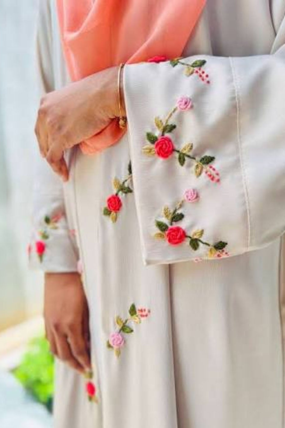 A close-up of a person wearing a light beige or cream-colored long-sleeved garment with colorful floral embroidery on the sleeve and hem, paired with a peach-pink headscarf.