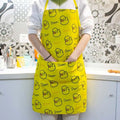 A person wearing a bright yellow kitchen apron with a repeating pattern of cartoon yellow ducks and the name 'Shawn' printed in black, standing in a kitchen with patterned tile backsplash.