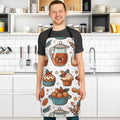 A man wearing a black t-shirt and a white apron with a colorful autumn-themed pattern of teapots, cupcakes, and muffins, standing in a bright, modern kitchen.