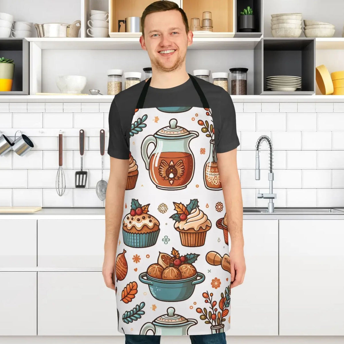 A man wearing a black t-shirt and a white apron with a colorful autumn-themed pattern of teapots, cupcakes, and muffins, standing in a bright, modern kitchen.