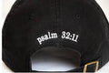 Close-up of the back of a black baseball cap with white embroidered text reading 'psalm 32:11' and a small gold-tone buckle visible on the strap.