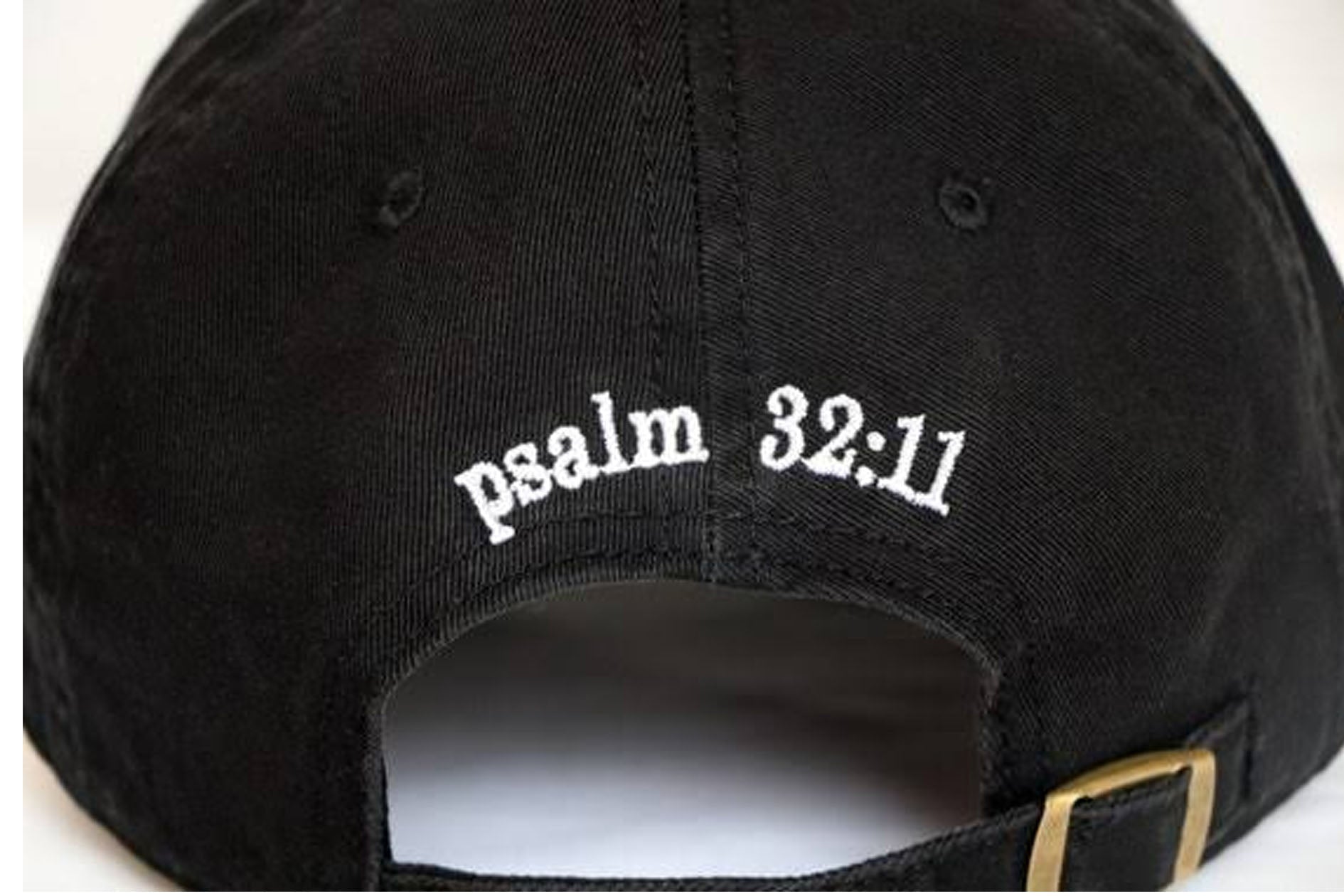 Close-up of the back of a black baseball cap with white embroidered text reading 'psalm 32:11' and a small gold-tone buckle visible on the strap.