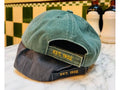 Two baseball caps stacked on a marble countertop in front of a green and cream checkered tile wall. The top cap is sage green canvas with yellow embroidered 'EST.