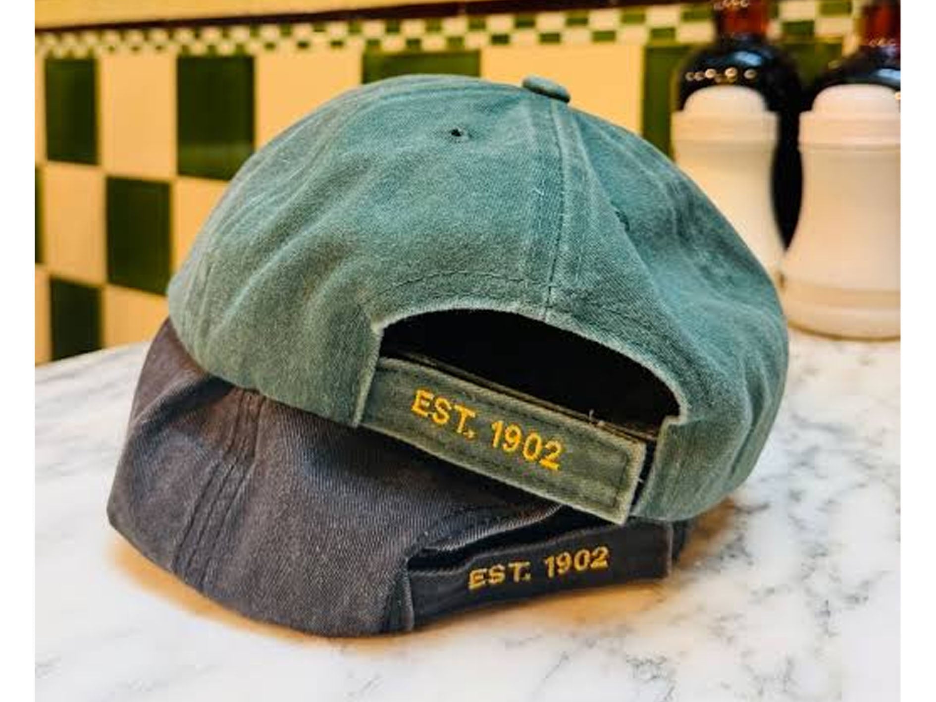 Two baseball caps stacked on a marble countertop in front of a green and cream checkered tile wall. The top cap is sage green canvas with yellow embroidered 'EST.