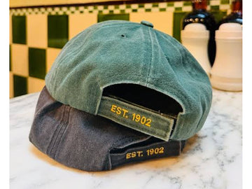 Two baseball caps stacked on a marble countertop in front of a green and cream checkered tile wall. The top cap is sage green canvas with yellow embroidered 'EST.