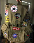 A khaki canvas backpack decorated with multiple embroidered patches, including Converse All Star, The Clash, NASA, and a Pepsi logo patch, held up by a hand in an indoor setting.
