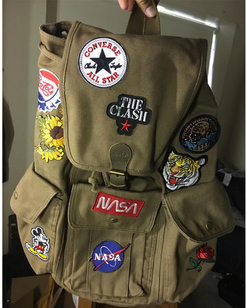 A khaki canvas backpack decorated with multiple embroidered patches, including Converse All Star, The Clash, NASA, and a Pepsi logo patch, held up by a hand in an indoor setting.