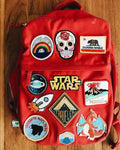 A red backpack with multiple colorful patches and stickers, including a Star Wars patch, laid flat on a wooden surface.