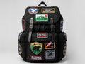 A black backpack covered with multiple colorful embroidered patches and badges, photographed against a light gray studio background.