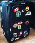 A black soft-sided rolling suitcase covered with numerous colorful travel stickers, photographed standing on a carpeted floor.