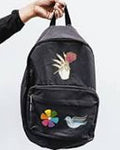 A black backpack with embroidered patches, held by a hand against a light gray background. The backpack features a white hand patch with a pink flower, a colorful flower, and a white dove.