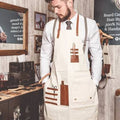 A man in a barbershop setting wearing a cream-colored apron with brown leather-like straps and pockets, over a white shirt and tie.