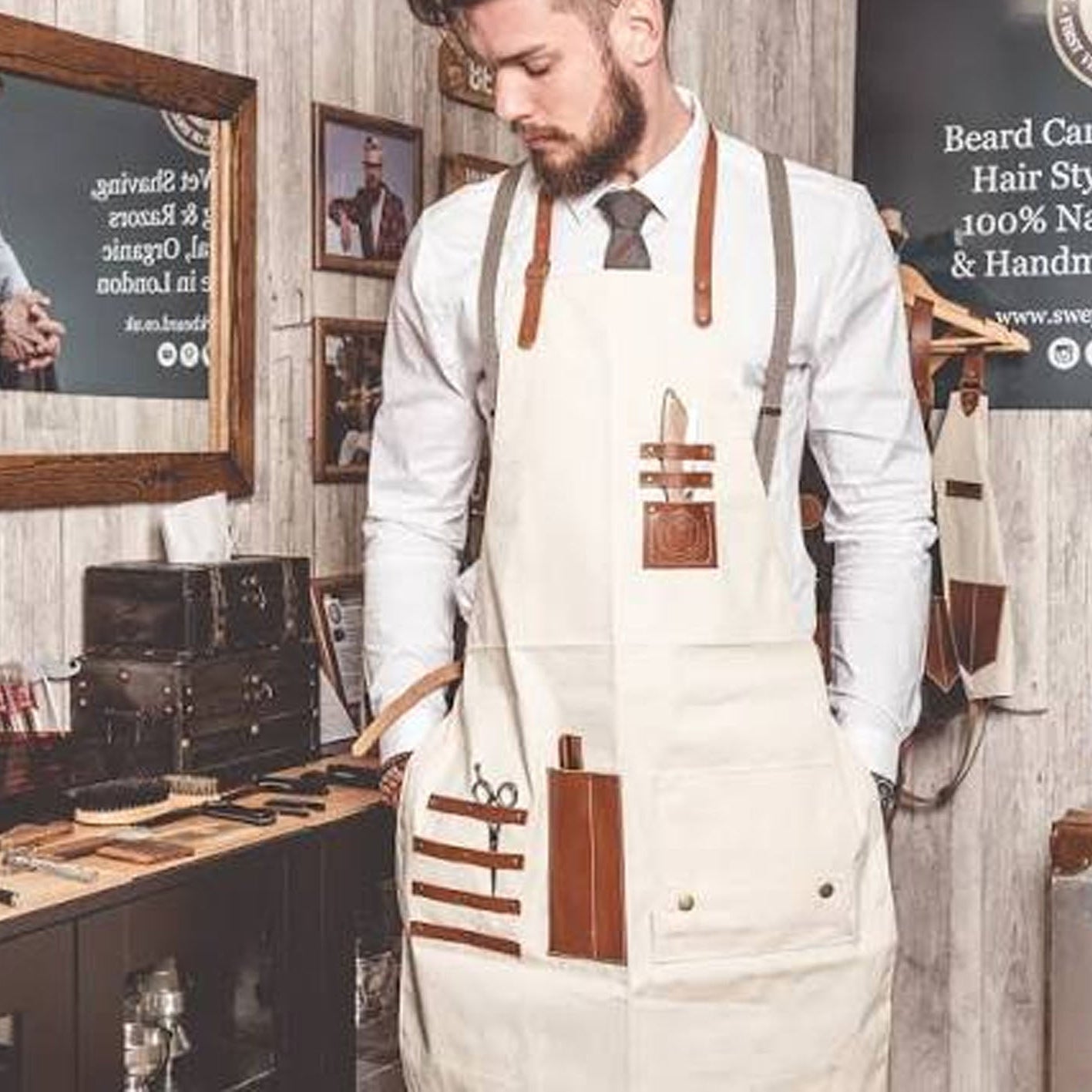 A man in a barbershop setting wearing a cream-colored apron with brown leather-like straps and pockets, over a white shirt and tie.