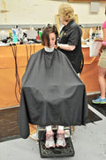 A person seated in a salon chair wearing a black barber cape while a stylist cuts their hair in a salon setting.