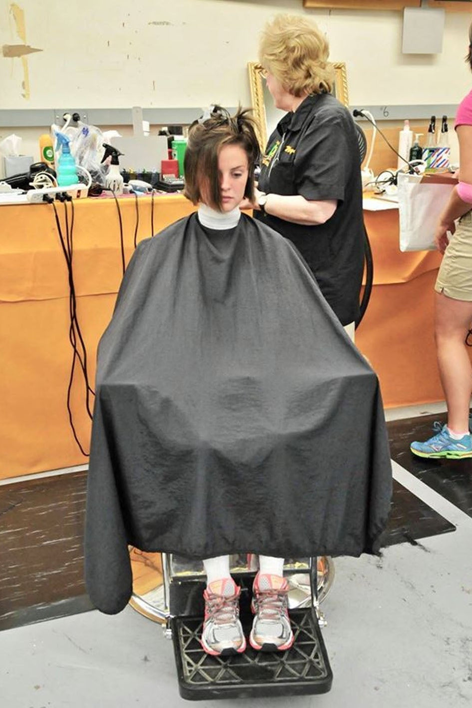 A person seated in a salon chair wearing a black barber cape while a stylist cuts their hair in a salon setting.
