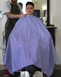 A person seated in a barber chair wearing a blue and white vertically striped barber cape, with a barber standing behind them in a barbershop setting.