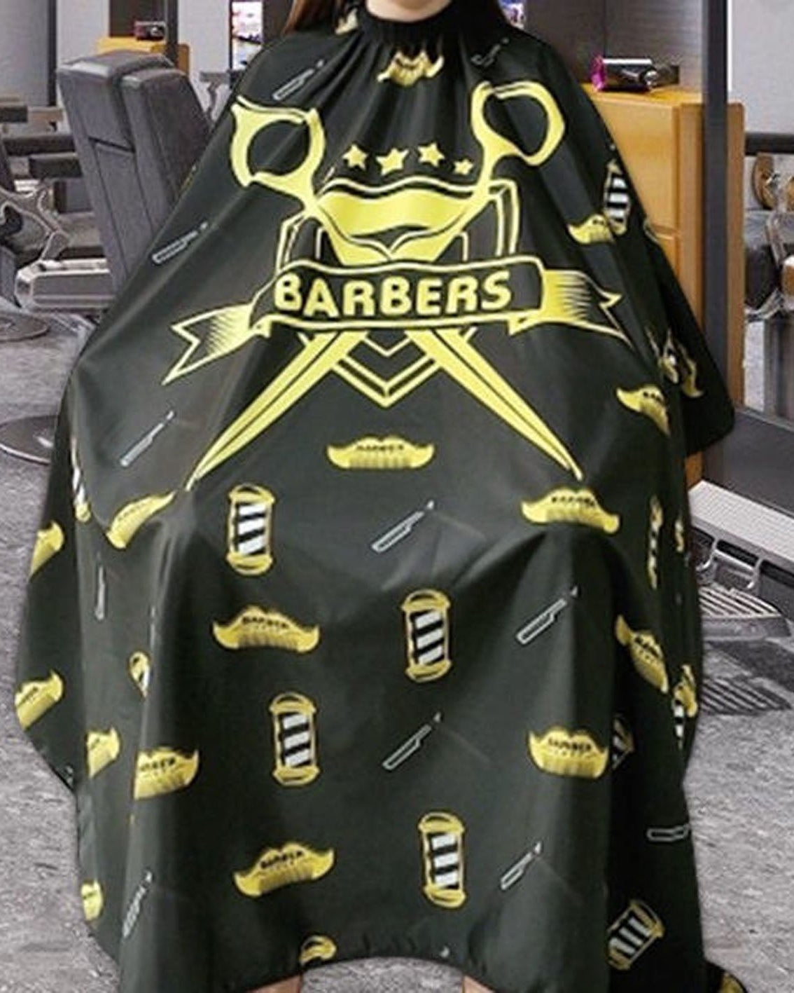 A black barber cape with a yellow 'BARBERS' graphic and barber tools pattern, worn by a person in a barbershop.
