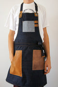 A person wearing a dark indigo denim apron with tan leather accents, including a chest pocket, waist tie, and two large front pockets, over a white t-shirt against a plain light gray background.