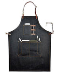 A dark denim apron with brown leather straps and trim, featuring two front utility pockets holding kitchen utensils, shown against a plain white background.