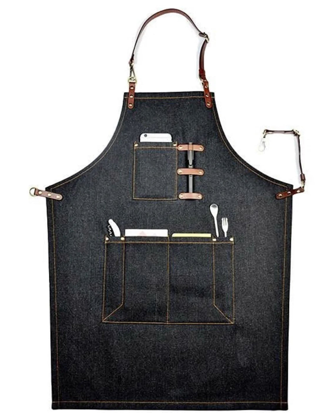 A dark denim apron with brown leather straps and trim, featuring two front utility pockets holding kitchen utensils, shown against a plain white background.