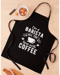 A black kitchen apron with white text reading 'I'm a BARISTA ALL THE time with COFFEE' and a coffee cup graphic, laid flat on a light wood surface with small food props nearby.