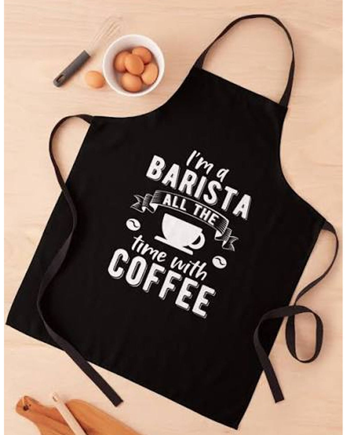 A black kitchen apron with white text reading 'I'm a BARISTA ALL THE time with COFFEE' and a coffee cup graphic, laid flat on a light wood surface with small food props nearby.