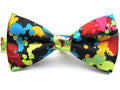 A pre-tied bow tie featuring a vibrant multicolor paint splatter pattern on a black satin background, displayed against a plain white background.