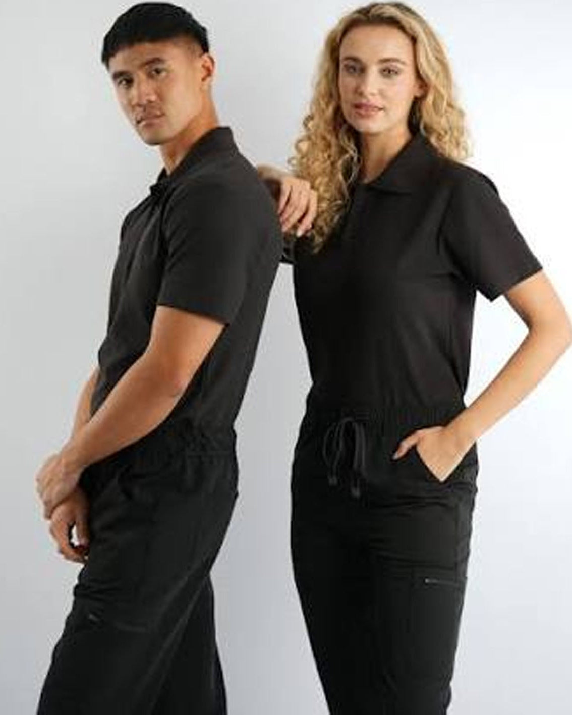 A man and a woman modeling matching black scrub sets against a light gray studio background. The man wears a short-sleeve scrub polo with matching scrub pants, and the woman wears a short-sleeve scrub top with matching scrub pants.