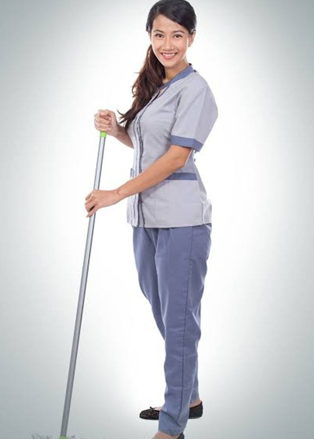 A woman wearing a light gray and blue scrub-style uniform stands holding a long-handled cleaning tool against a plain light gray studio background.
