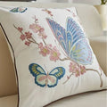 An off-white decorative throw pillow with embroidered butterflies and pink cherry blossoms, resting on a beige sofa.