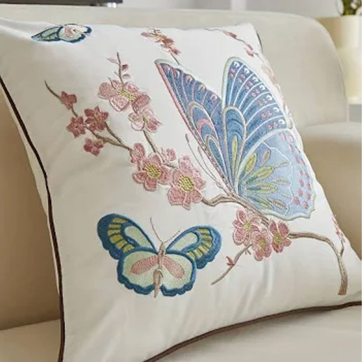 An off-white decorative throw pillow with embroidered butterflies and pink cherry blossoms, resting on a beige sofa.