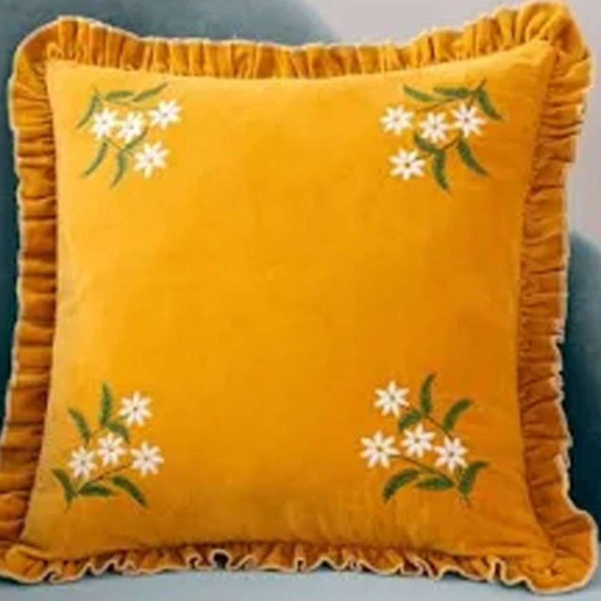 A square mustard yellow velvet throw pillow with a ruffled edge and embroidered white flowers with green leaves in each corner, shown resting on a blue-gray chair.