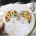 Embroidery hoop with two women's faces embroidered on a white background, each wearing a floral headpiece.