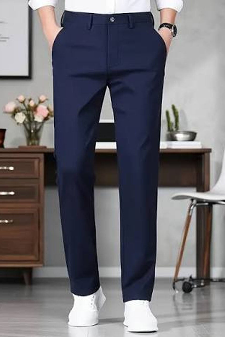A person wearing navy blue dress pants, a white shirt, and white sneakers, standing in a modern office setting with a desk, chair, and wooden cabinet in the background.