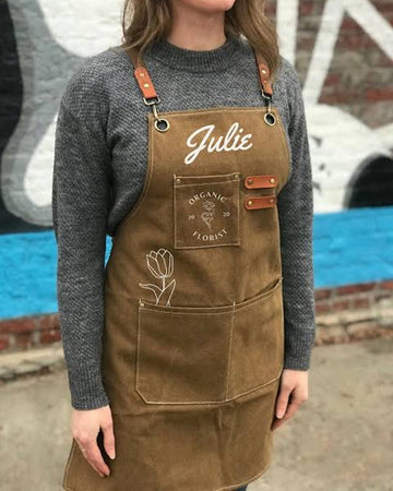 A person wearing a brown canvas apron with adjustable brown leather straps, featuring a front pocket with 'Julie' and 'Organic Flowers' embroidered on it, over a gray knit sweater.