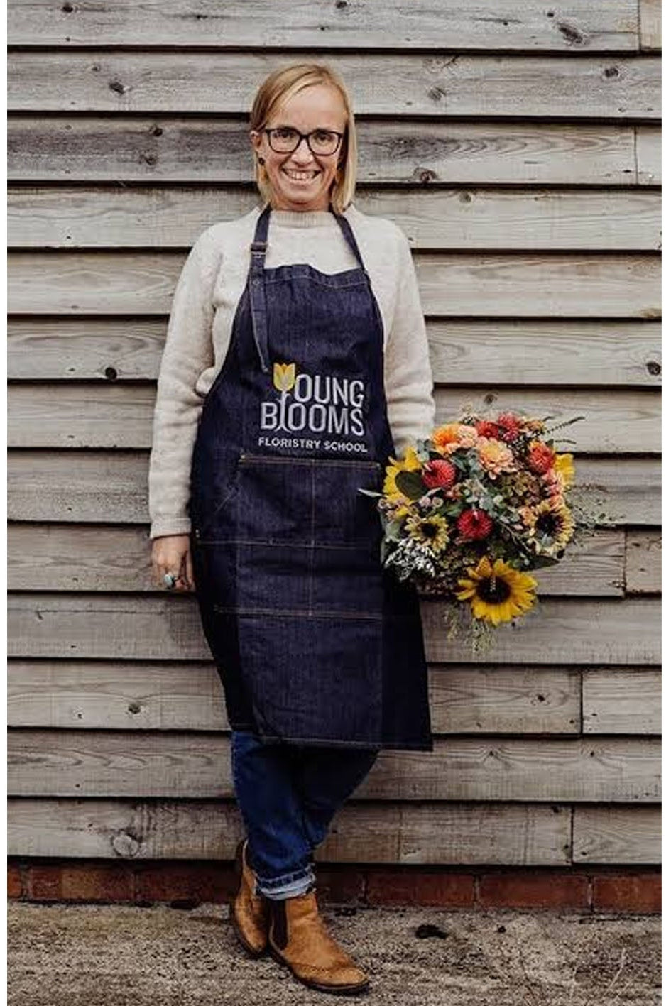 A woman wearing a dark navy apron with 'Young Blooms' printed on the chest stands in front of a weathered wooden wall, holding a colorful bouquet of sunflowers and other flowers.