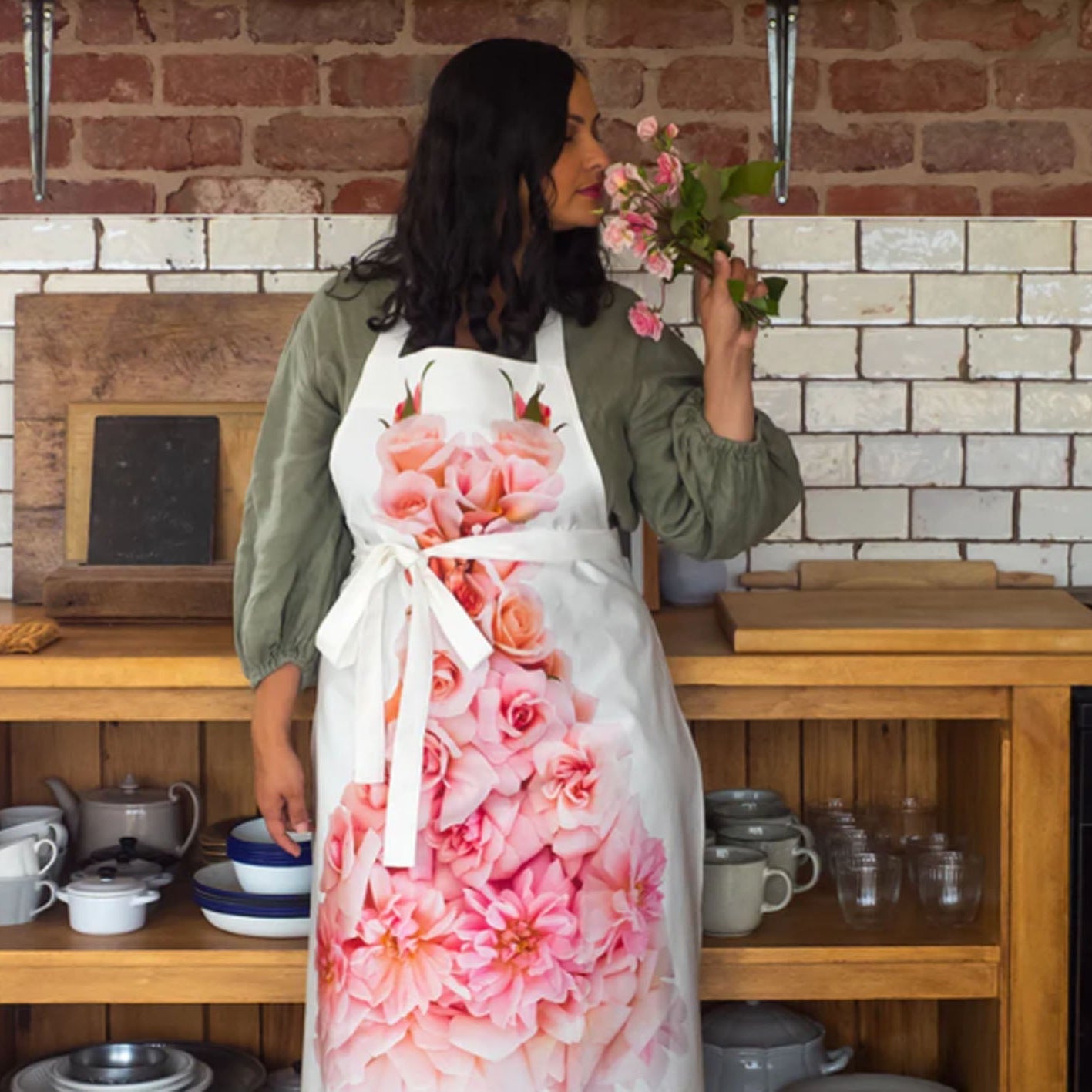A woman wearing a white apron with a large pink rose print stands in a rustic kitchen holding a bouquet of pink flowers. The kitchen features a brick and white tiled backsplash, wooden cabinets, and various ceramic dishes.