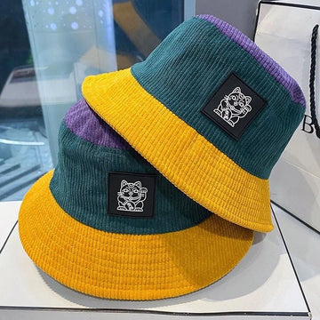 Two corduroy bucket hats stacked on a white surface. The hats feature a yellow brim, a dark teal crown, and a purple inner lining, with a black square patch displaying a white cat illustration on the side.