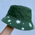 An olive green bucket hat with a white daisy pattern on the brim, held by a person's hand against a light blue textured wall.