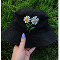 A black bucket hat with embroidered white daisies featuring yellow centers and green stems, held by a hand with purple nail polish against a background of green grass.